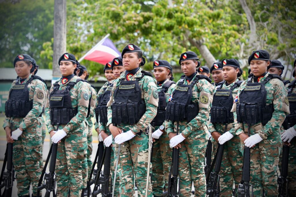 Mujeres soldado uniformadas en formación, bandera dominicana detrás.
