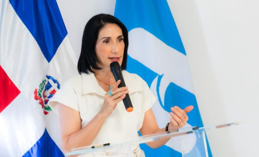 Mujer hablando en un micrófono, bandera detrás.