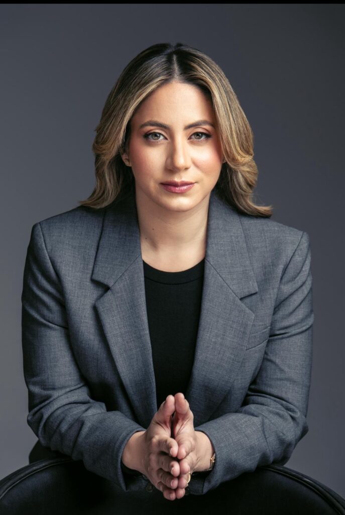 Persona en traje gris, manos juntas, fondo oscuro.