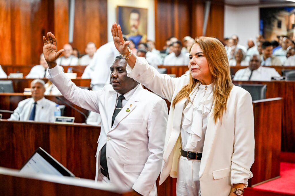 Personas en sala vestidos de blanco, levantando la mano.